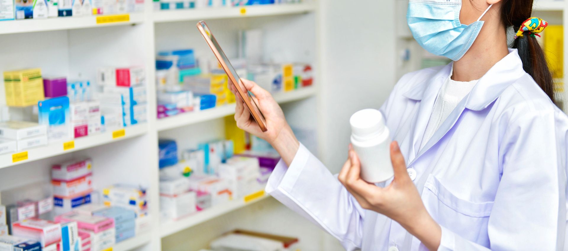 Masked pharmacist holding a medicine bottle and tablet in a pharmacy