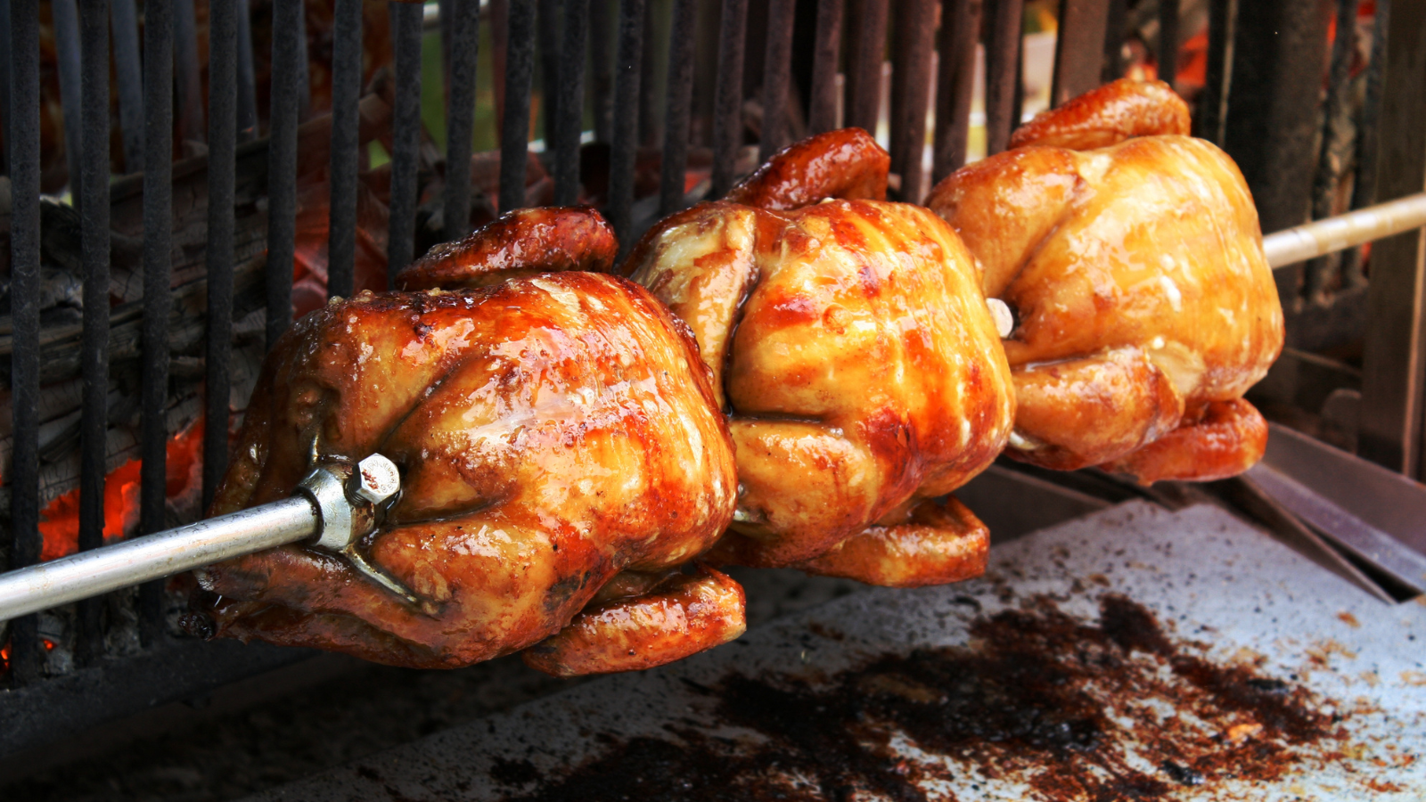 Whole roast chickens on a rotisserie spit turning over an open flame grill