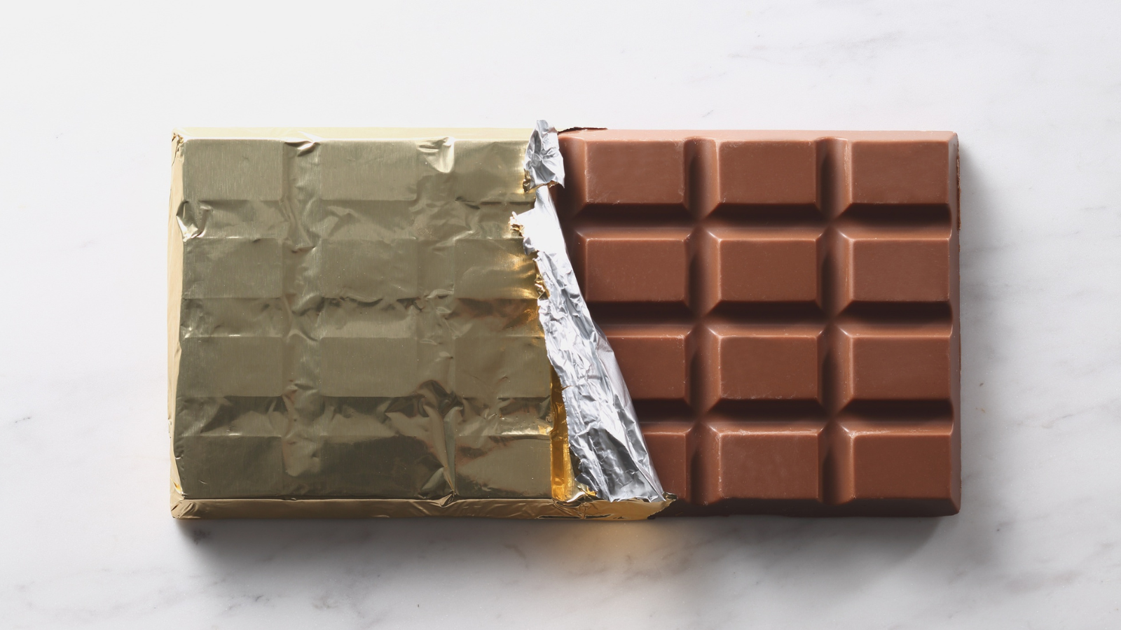 Chocolate bar half-unwrapped from gold foil on a white marble surface.